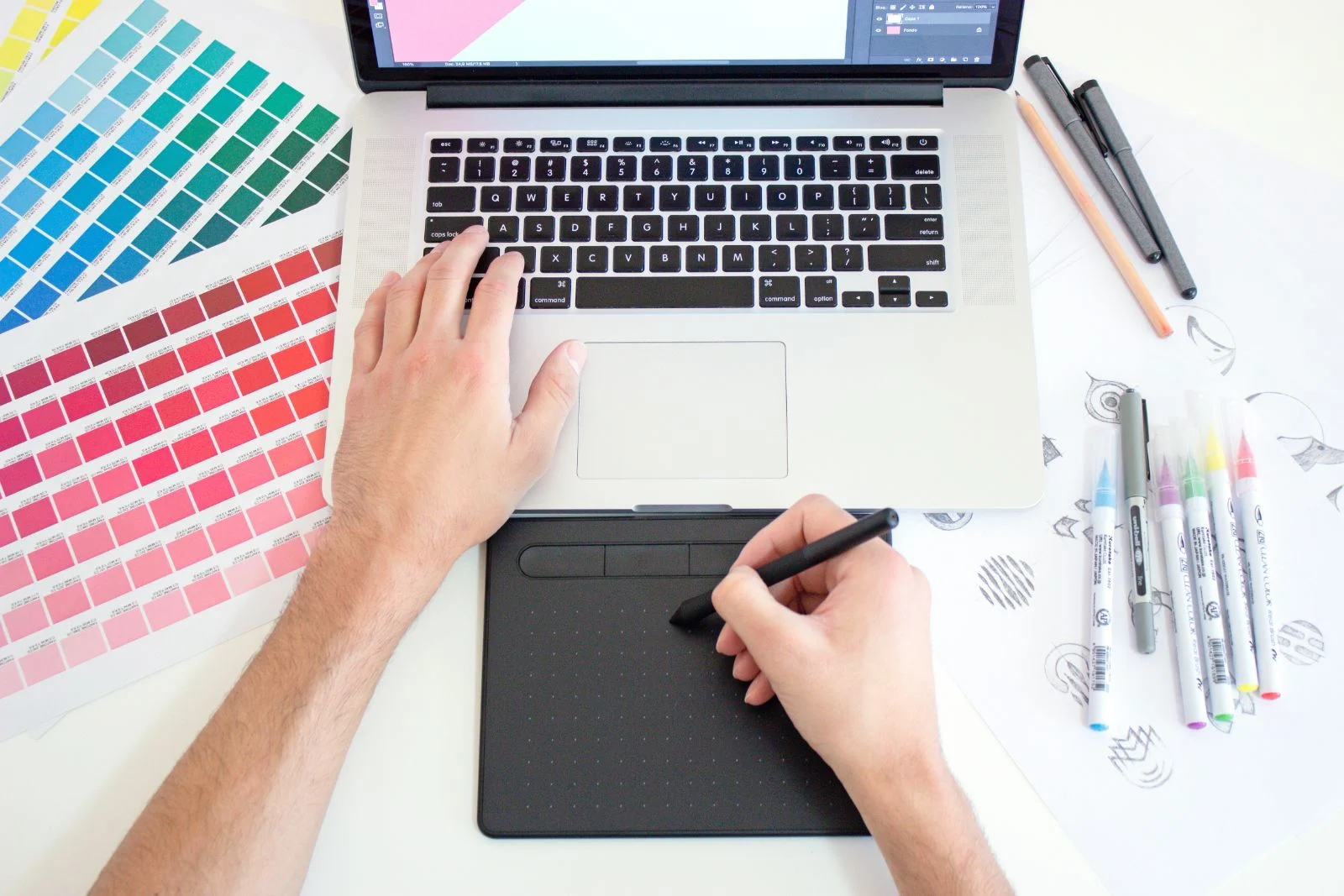 A designer sketching logo ideas on a drawing tablet next to printed color swatches